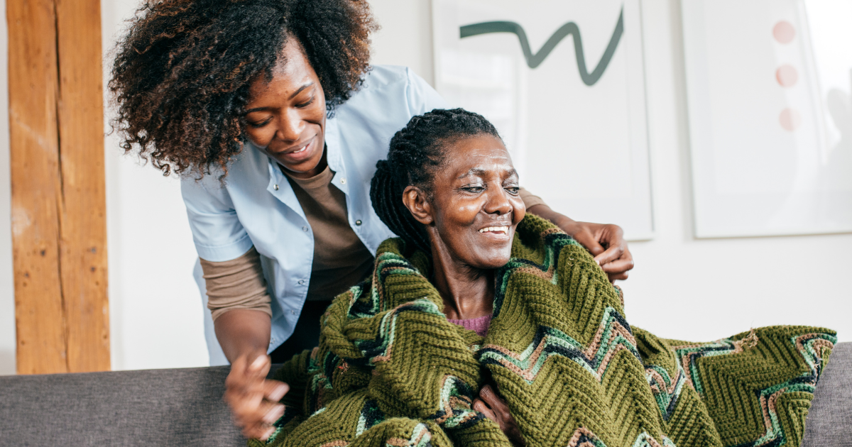 A caregiver gently wraps a blanket around an elderly woman seated on a couch in a home setting.
