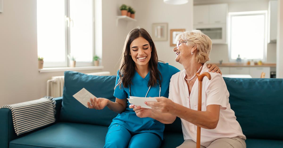 A caregiver and an elderly woman sit together on a couch reviewing papers and smiling.