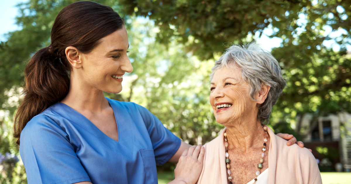 A caregiver and an elderly woman smile at each other while standing outside in a garden or park.