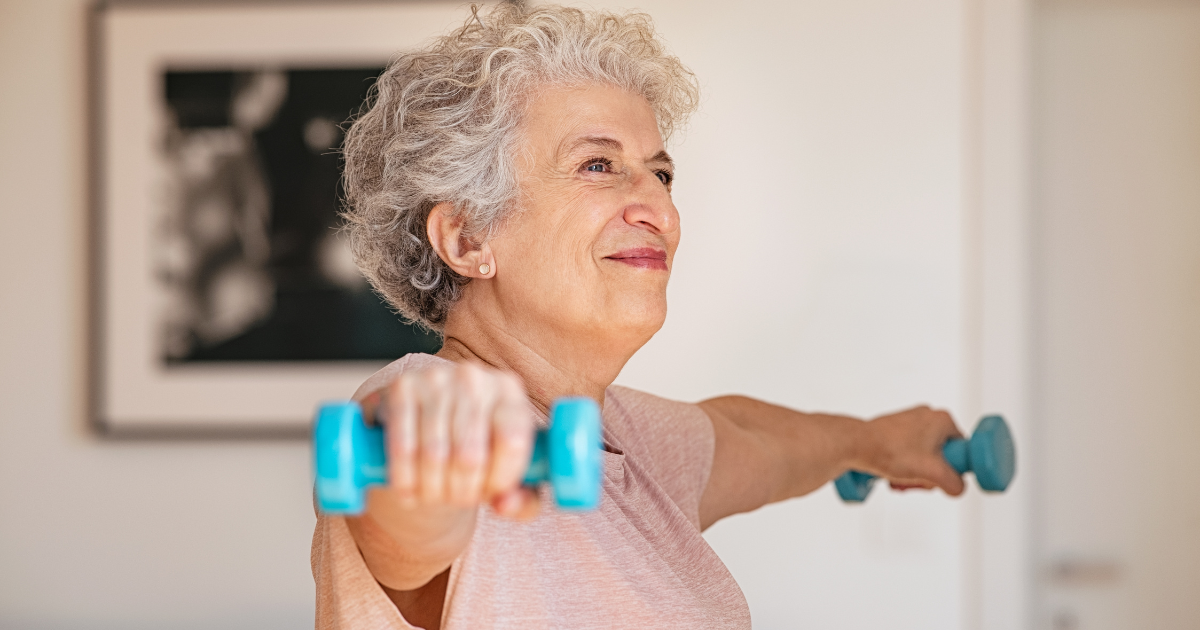 A senior woman lifting light dumbbells with her arms outstretched while participating in an indoor exercise routine.