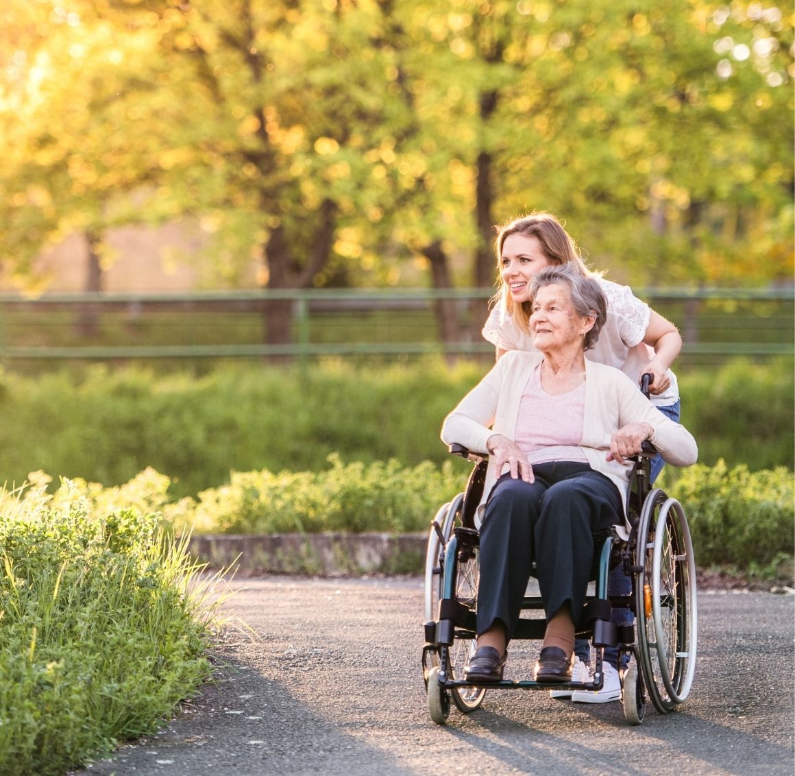 Female caregiver happily pushing an elderly woman in a wheelchair outside along a path, supporting mobility and daily home care needs.