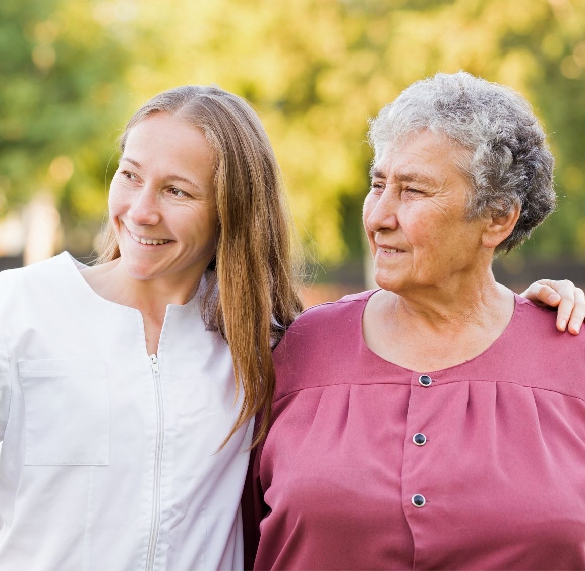 Caregiver walking beside an elderly woman outdoors while offering gentle support and companionship.