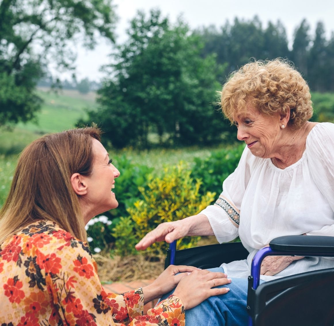 Caregiver kneeling beside an elderly woman in a wheelchair while talking and holding her hand outdoors.