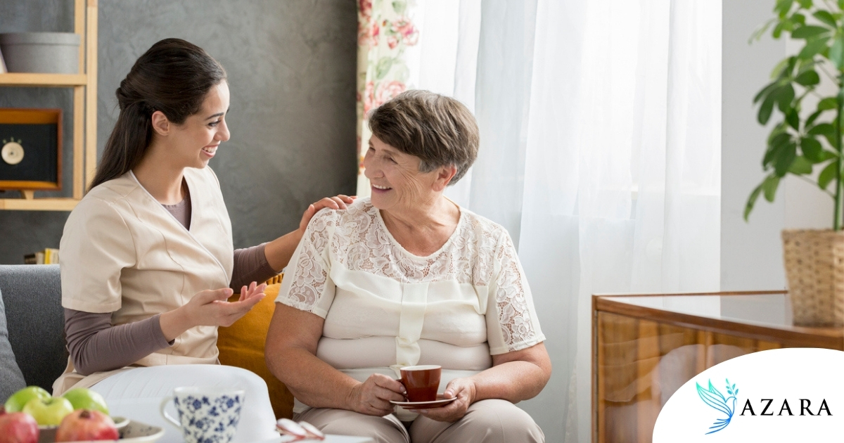 Smiling caregiver with her hand on an older woman’s shoulder as they share tea on the couch, showing home care vs assisted living